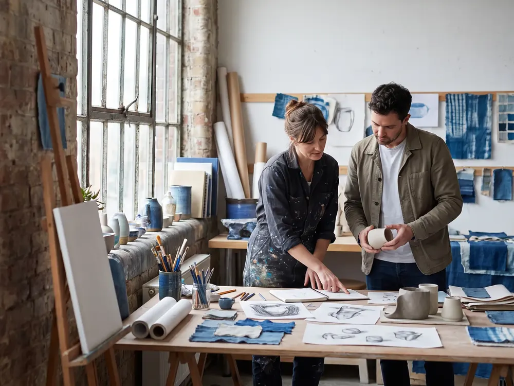 An independent artist reviewing original work with a business collaborator in a working studio filled with tools and materials